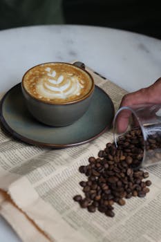 A cappuccino with latte art next to spilled coffee beans on a vintage newspaper.